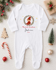 A white onesie with a Christmas-themed design, featuring a small elf or gnome, is displayed on a white background alongside various Christmas decorations and ornaments.