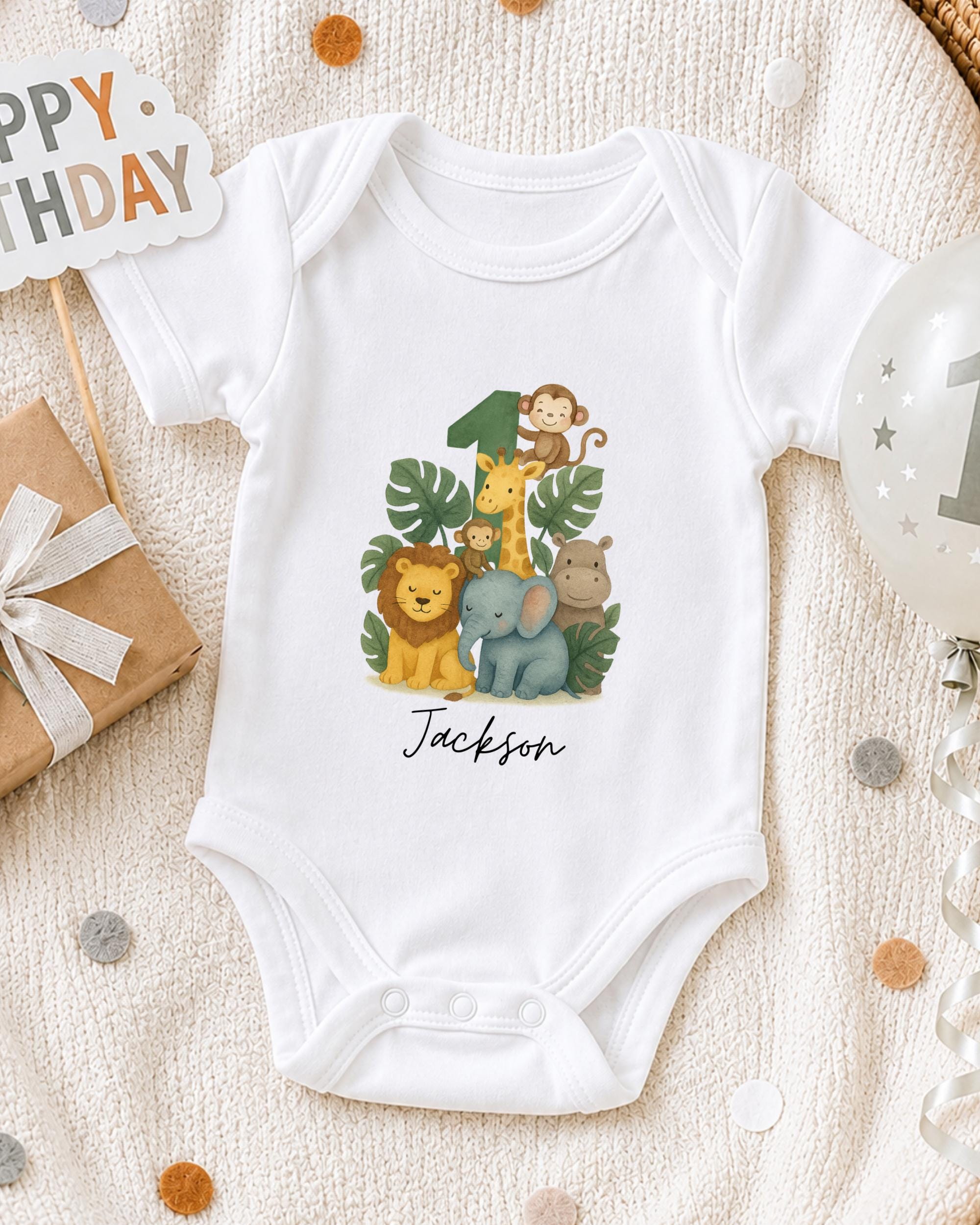 A white onesie with a jungle-themed design, featuring a monkey, giraffe, lion, and elephant, along with the name "Jackson" printed on it. The onesie is placed on a white blanket, surrounded by a gift box, a balloon, and confetti.