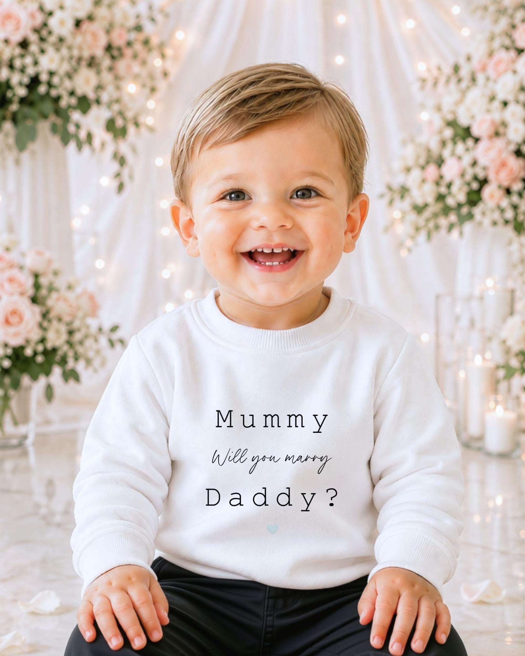 A smiling baby boy wearing a white sweatshirt with text on it, sitting in front of a floral background.
