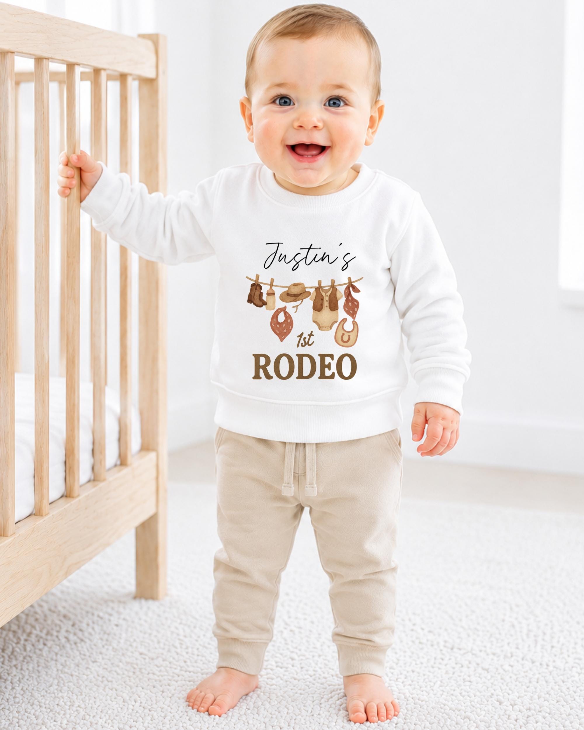 A smiling baby boy wearing a white sweatshirt with a graphic design and text, standing in front of a wooden crib.