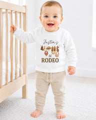 A smiling baby boy wearing a white sweatshirt with a graphic design and text, standing in front of a wooden crib.