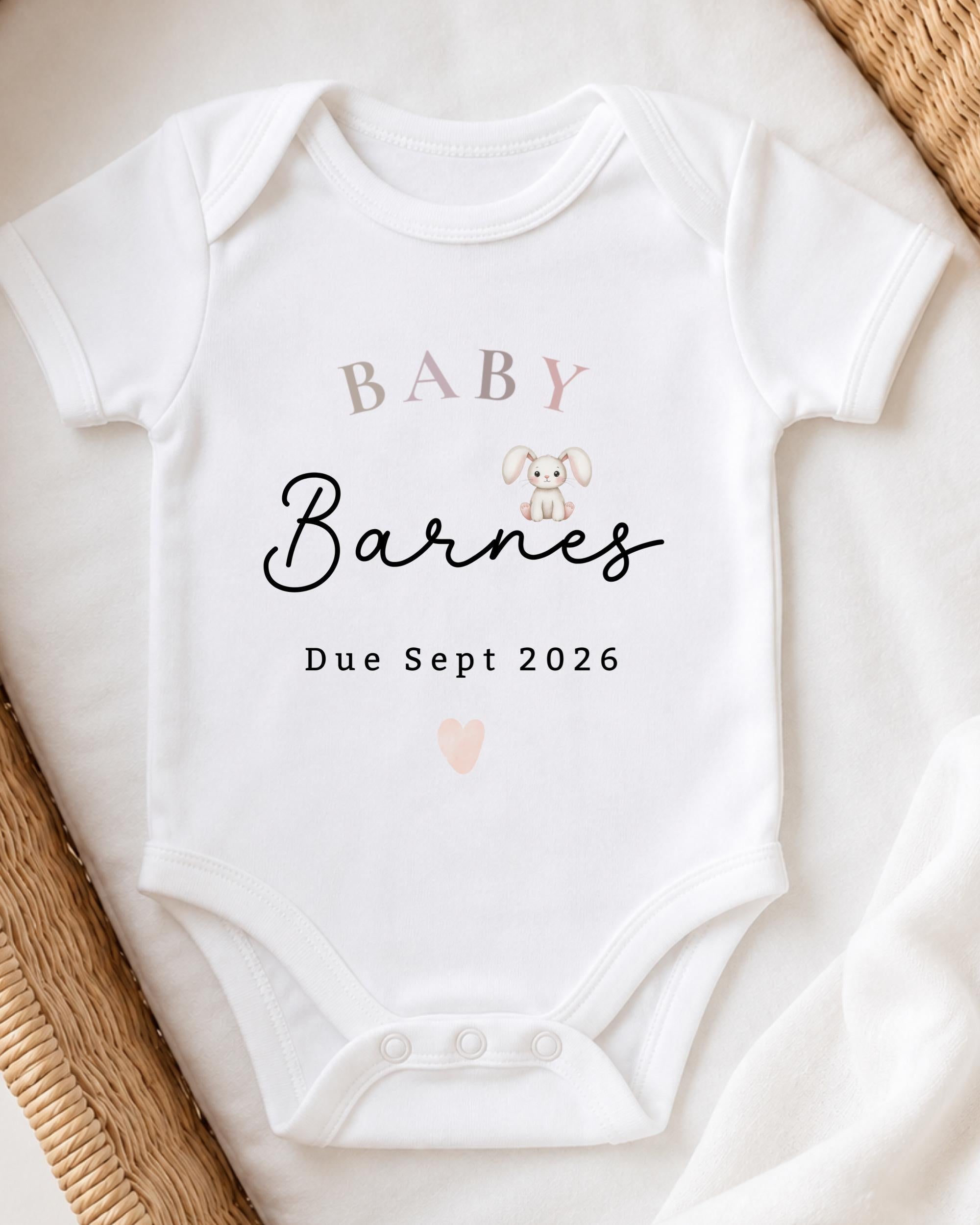 A white baby onesie with the text "BABY Barnes" printed on it, along with a small illustration of a rabbit. The text also includes "Due Sept 2026".