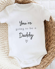 A white baby onesie with the text "You're going to be a Daddy" printed on it, placed on a woven basket.