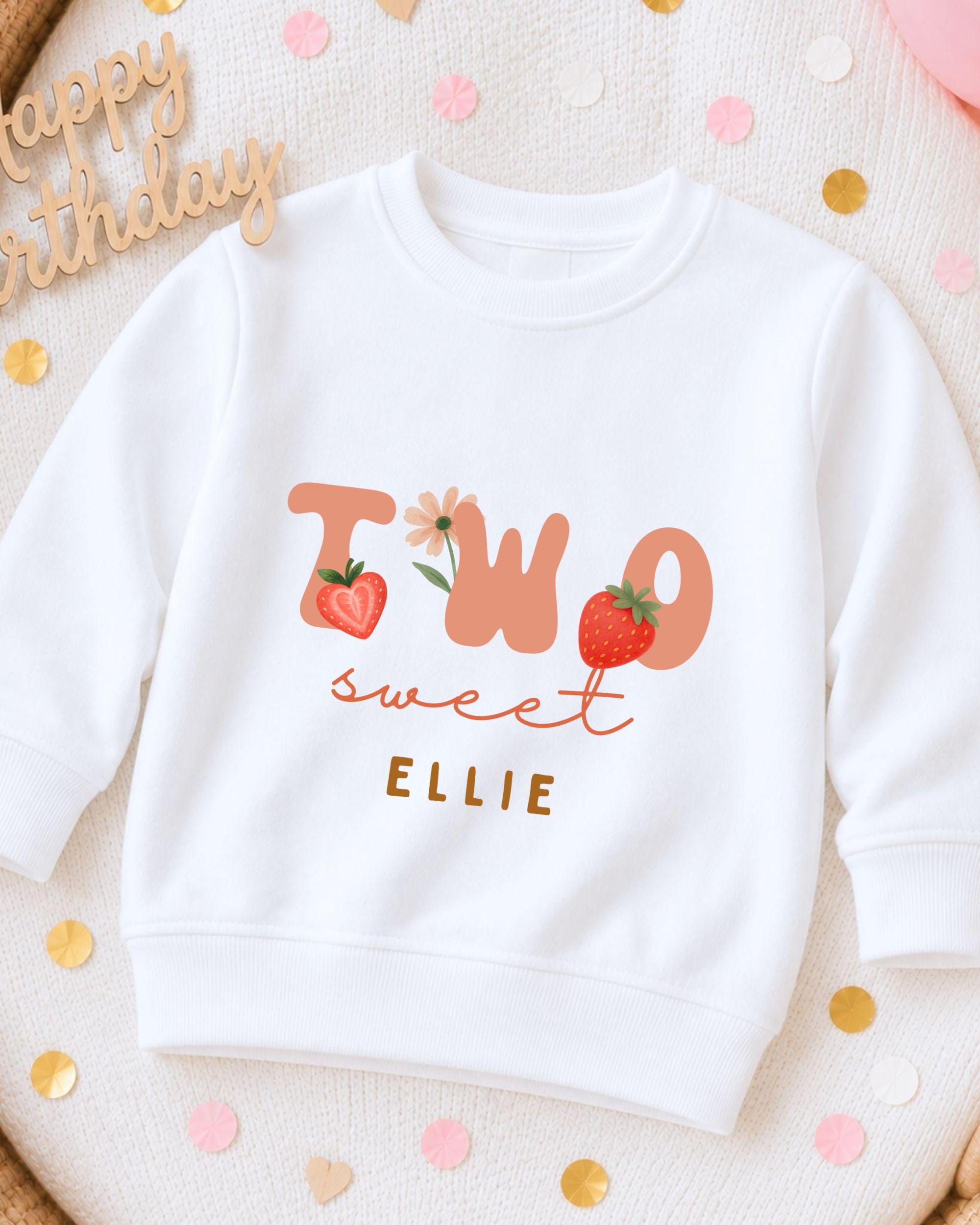 A white sweatshirt with the text "TWO sweet Ellie" printed on it, along with illustrations of strawberries and flowers.