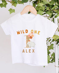 A white t-shirt with a printed design on it, hanging on a wooden hanger. The design features a white elephant surrounded by colorful balloons, with the text "WILD ONE ALEX" printed on the shirt.