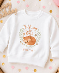 A white sweatshirt with a graphic design featuring a sleeping fox and the text "Halfway to One" and "Freya" printed on it.