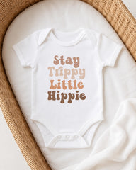 A white baby onesie with the text "Stay Trippy Little Hippie" printed on it, laid out on a white blanket in a wicker basket.