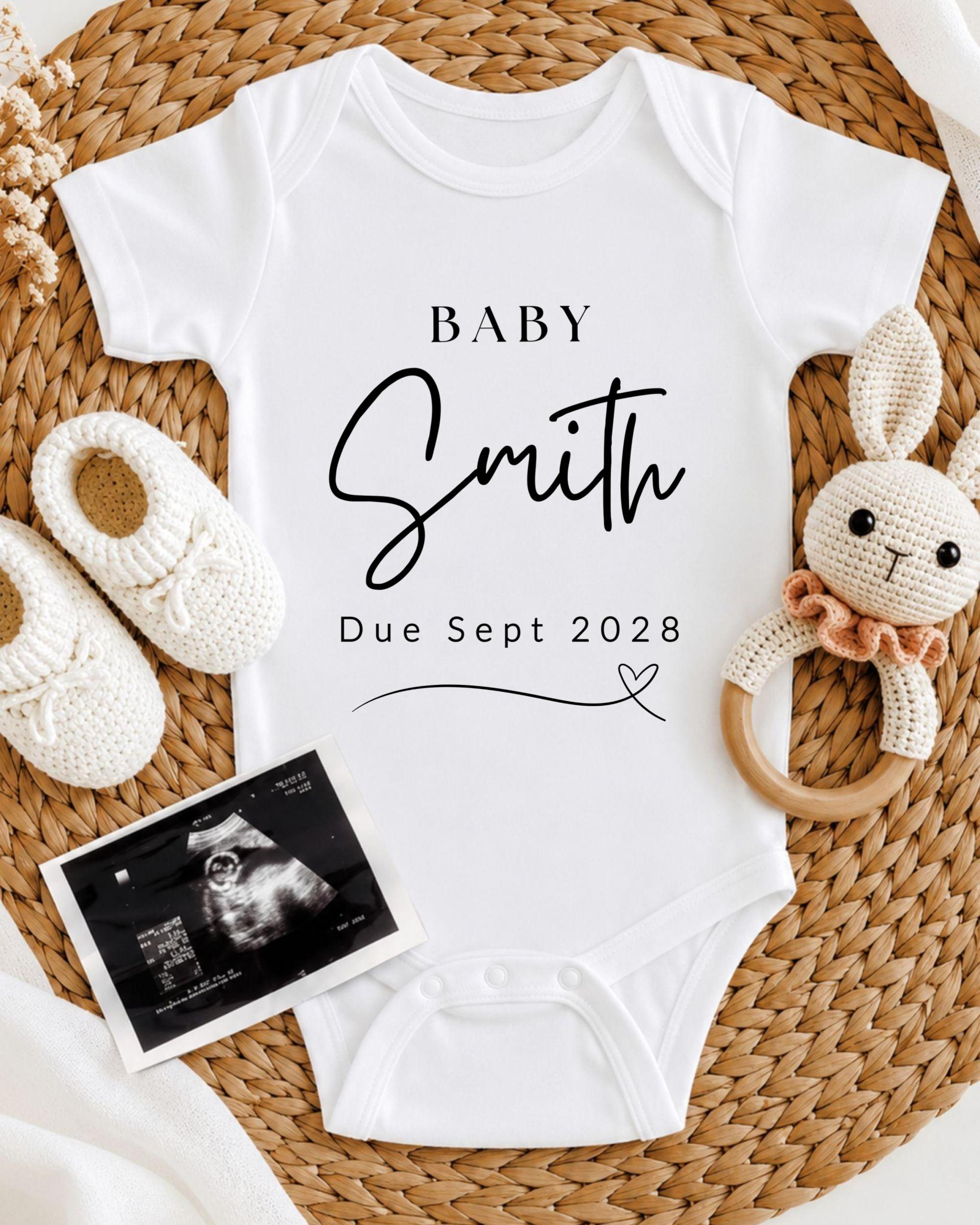 A white baby onesie with the text "BABY Smith Due Sept 2028" printed on it, along with a crocheted rabbit toy and a pair of white booties. There is also an Ultrasound image of a baby's belly.