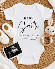 A white baby onesie with the text "BABY Smith Due Sept 2028" printed on it, along with a crocheted rabbit toy and a pair of white booties. There is also an Ultrasound image of a baby's belly.