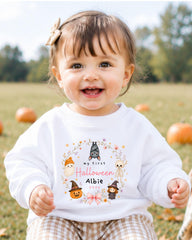 A smiling baby girl wearing a white sweater with a Halloween-themed design and text.