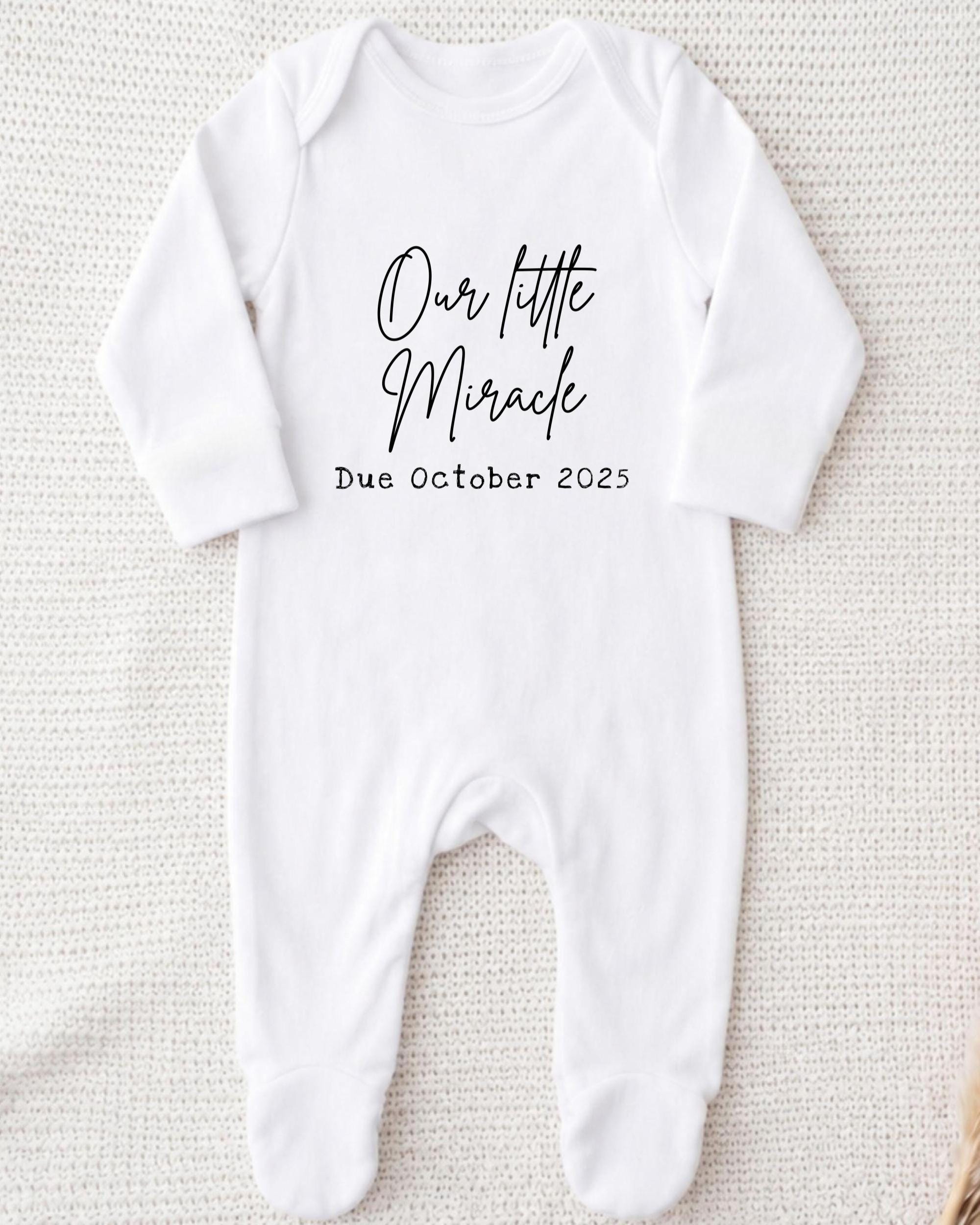 A white onesie with the text "Our little Miracle Due October 25" printed on it.