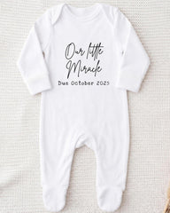 A white onesie with the text "Our little Miracle Due October 25" printed on it.
