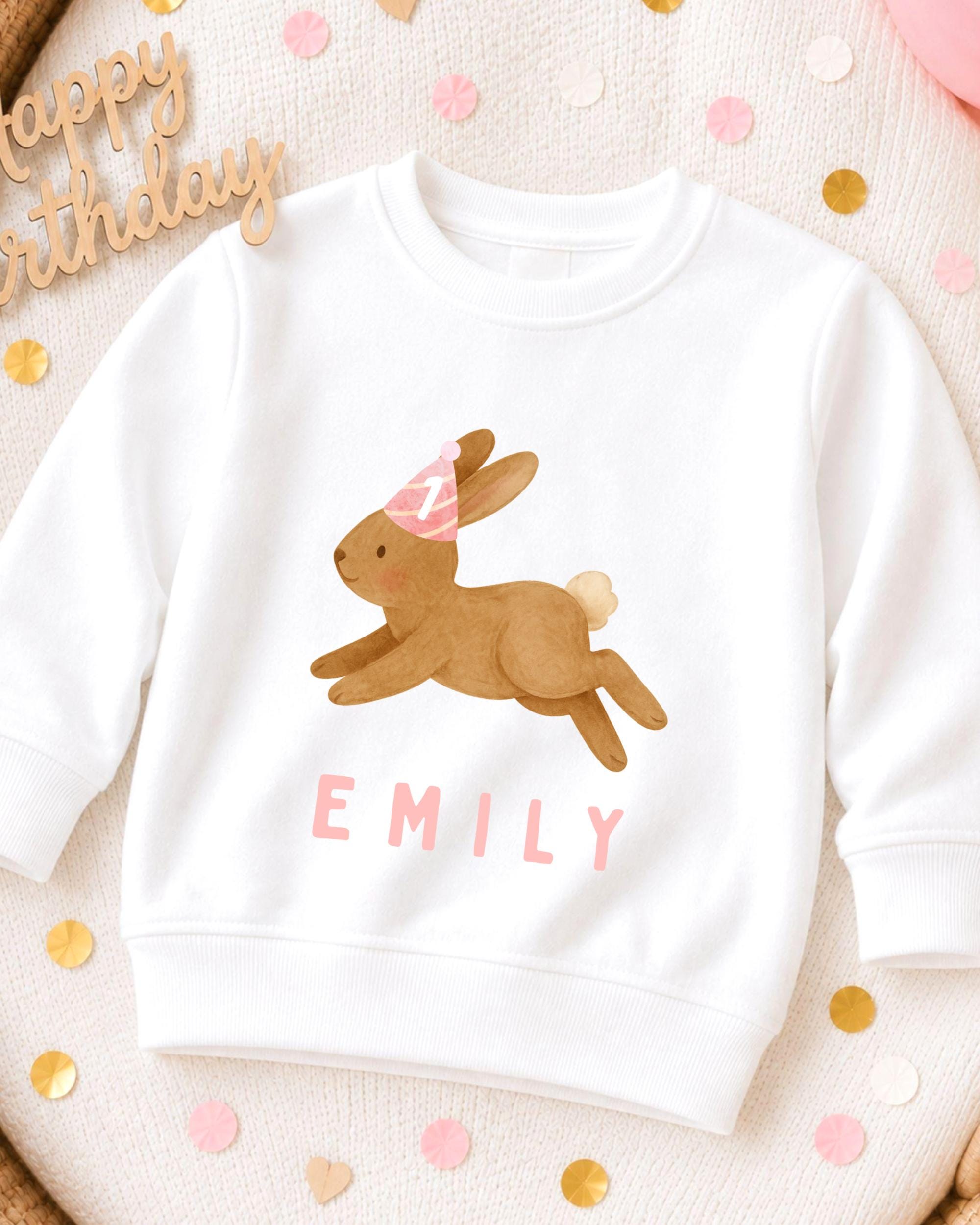 A white sweatshirt with a cartoon rabbit wearing a party hat and the name "EMILY" printed on it, surrounded by confetti.