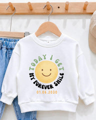 A white sweatshirt with a graphic design featuring a smiling yellow smiley face and the text "Today I get my forever smile" printed on it.
