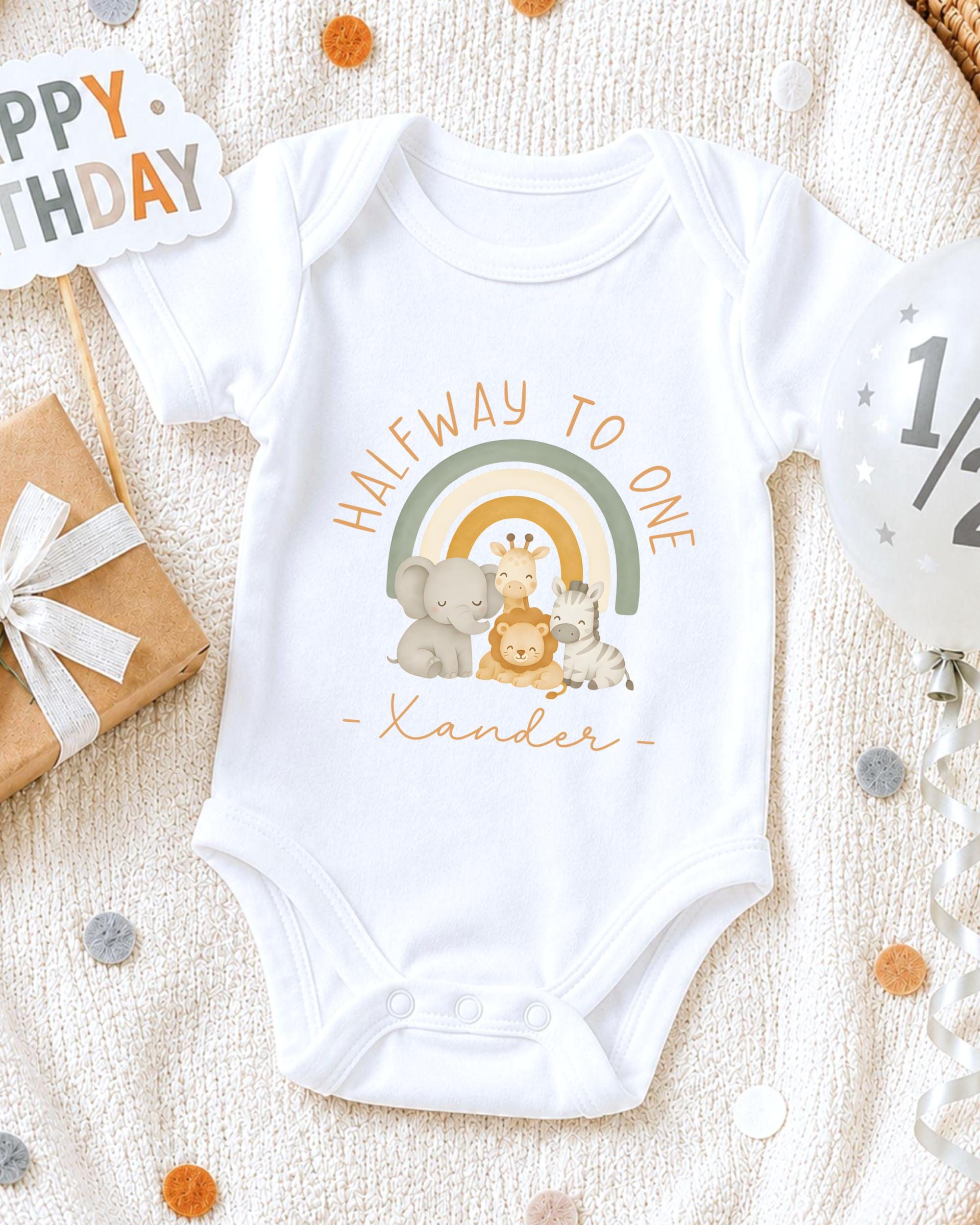 A white onesie with a cartoon illustration of animals, including a giraffe, elephant, and zebra, along with the text "Halfway to one" and "Xander" printed on it. The onesie is placed on a white blanket, surrounded by confetti and a birthday card.