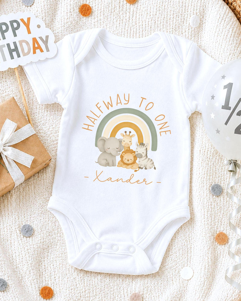 A white onesie with a cartoon illustration of animals, including a giraffe, elephant, and zebra, along with the text "Halfway to one" and "Xander" printed on it. The onesie is placed on a white blanket, surrounded by confetti and a birthday card.
