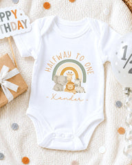 A white onesie with a cartoon illustration of animals, including a giraffe, elephant, and zebra, along with the text "Halfway to one" and "Xander" printed on it. The onesie is placed on a white blanket, surrounded by confetti and a birthday card.