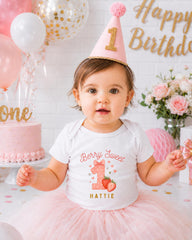A young girl wearing a white t-shirt with a strawberry design and a pink party hat sits in front of a birthday cake and balloons, with a "Happy Birthday" sign in the background.