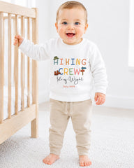 A smiling baby boy wearing a white sweatshirt with the text "HIKING CREW" and "Fleece of Wight" printed on it, standing in front of a wooden crib.