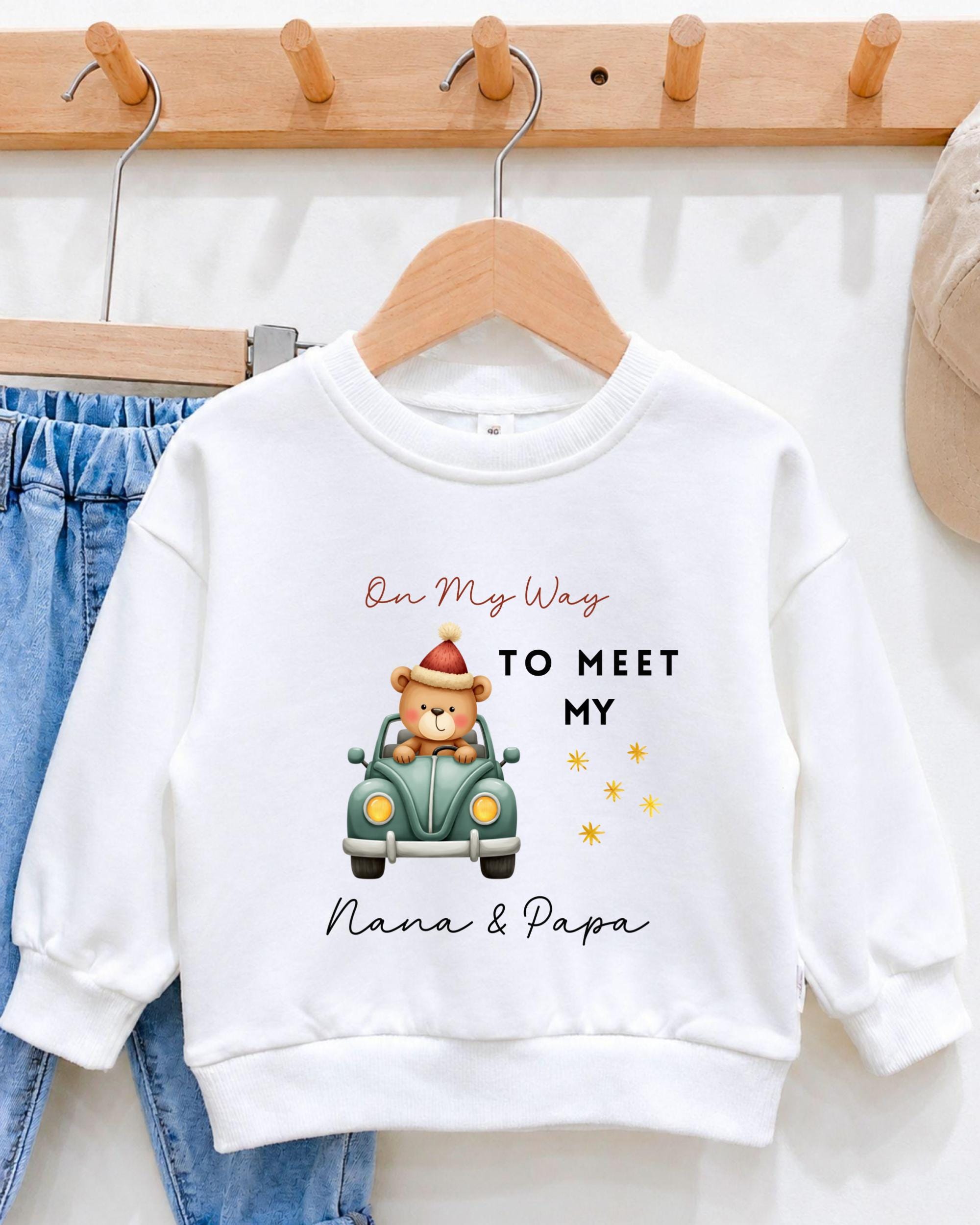 A white sweatshirt with a cartoon image of a bear in a car and text that says "On My Way to Meet My Nana & Papa".
