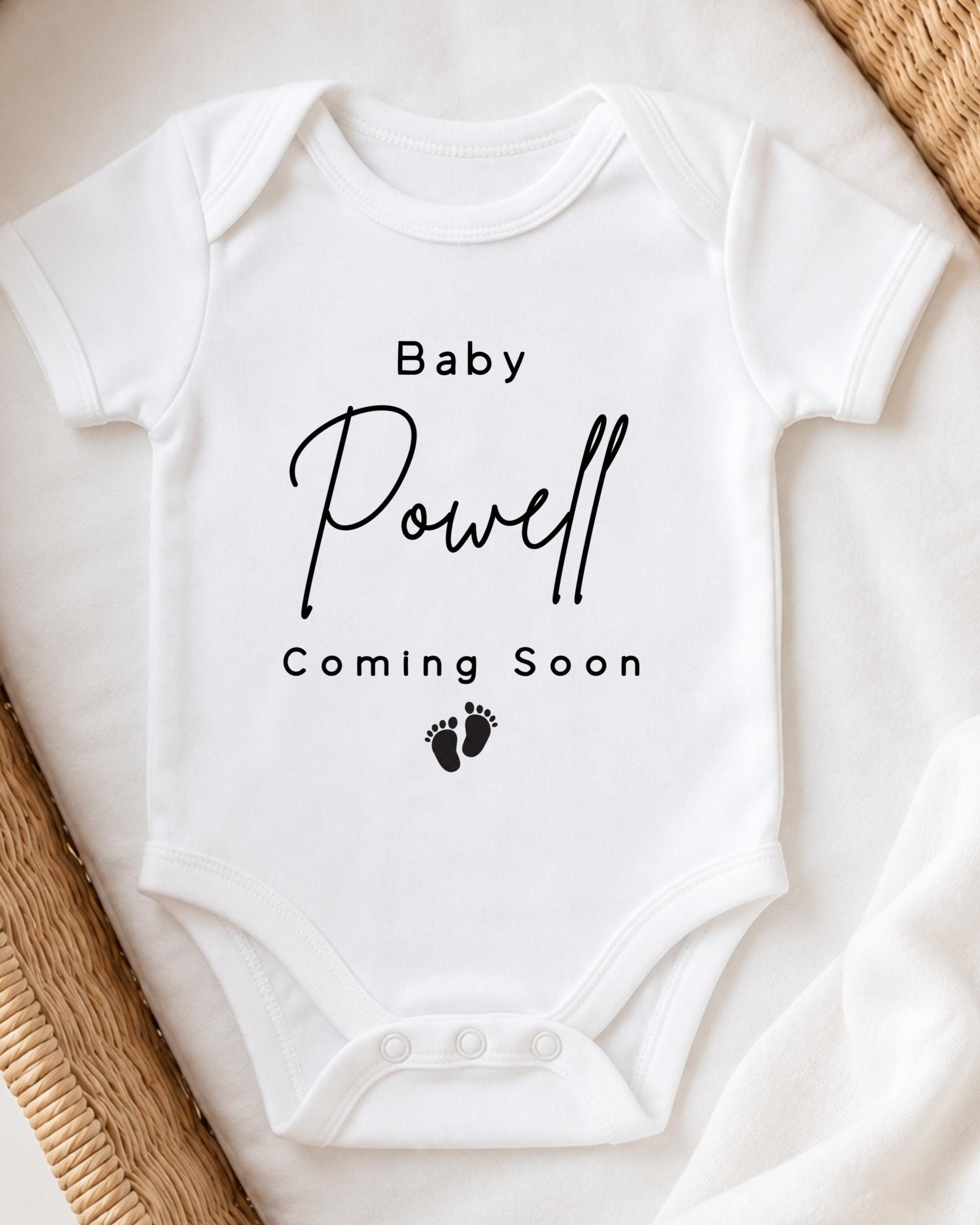 A white baby onesie with the text "Baby Powell Coming Soon" printed on it.