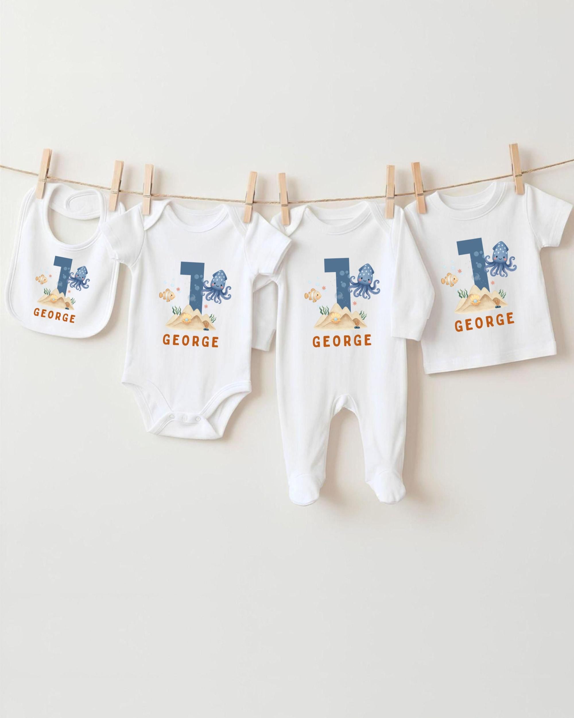 A collection of white baby clothes, including onesies and bibs, with the name "GEORGE" printed on them. The clothes are hanging on a clothesline, with the first two items being larger than the others.