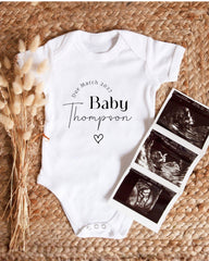 A white baby onesie with the text "Die March 2027 Baby Thompson" printed on it, along with a heart symbol. The onesie is placed next to a folded photograph of an ultrasound image of a baby.