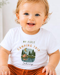 A smiling baby wearing a white t-shirt with a camping-themed graphic and text.