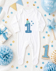 A white onesie with the number 1 and the name "Noah" printed on it, surrounded by birthday decorations such as a blue balloon, blue and gold confetti, and a blue and gold star.