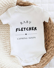 A white baby onesie with the text "BABY FLETCHER COMING SOON" printed on it, placed on a woven basket.