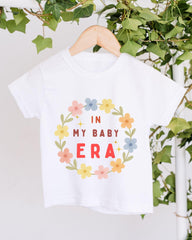 A white t-shirt with a floral wreath design and text printed on it, hanging on a wooden hanger.