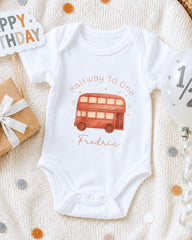 A white onesie with a red double-decker bus design, surrounded by confetti, a birthday card, and a balloon.