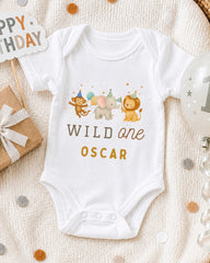 A white onesie with a cartoon image of a monkey, elephant, and lion, along with the text "WILD one OSCAR" printed on it, is displayed on a bed with confetti.