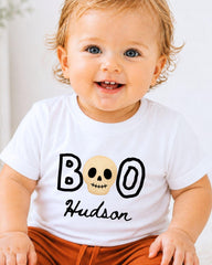 A smiling baby wearing a white t-shirt with a skull design and the text "BOO Hudson" printed on it.