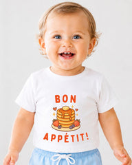 A smiling baby wearing a white t-shirt with a pancake and fruit graphic and the text " bon appétit!"