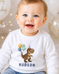 A smiling baby boy wearing a white shirt with a dinosaur and balloons printed on it.