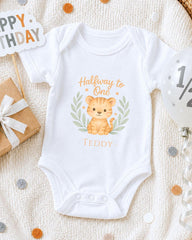 A white onesie with a cartoon tiger graphic and the text "Halfway to One Teddy" printed on it, along with a "Happy Birthday" sign and confetti.
