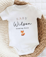 A white baby onesie with the text "BABY WILSON Coming Soon" printed on it, along with a small illustration of a fox.