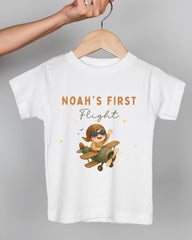 A white t-shirt with a cartoon image of a child wearing a pilot's hat and flying a small airplane, along with the text "Noah's first flight" printed on it.
