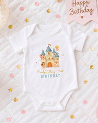 A white onesie with a castle design and the text "Emilly's Fairy First Birthday" printed on it, along with confetti and a birthday banner.