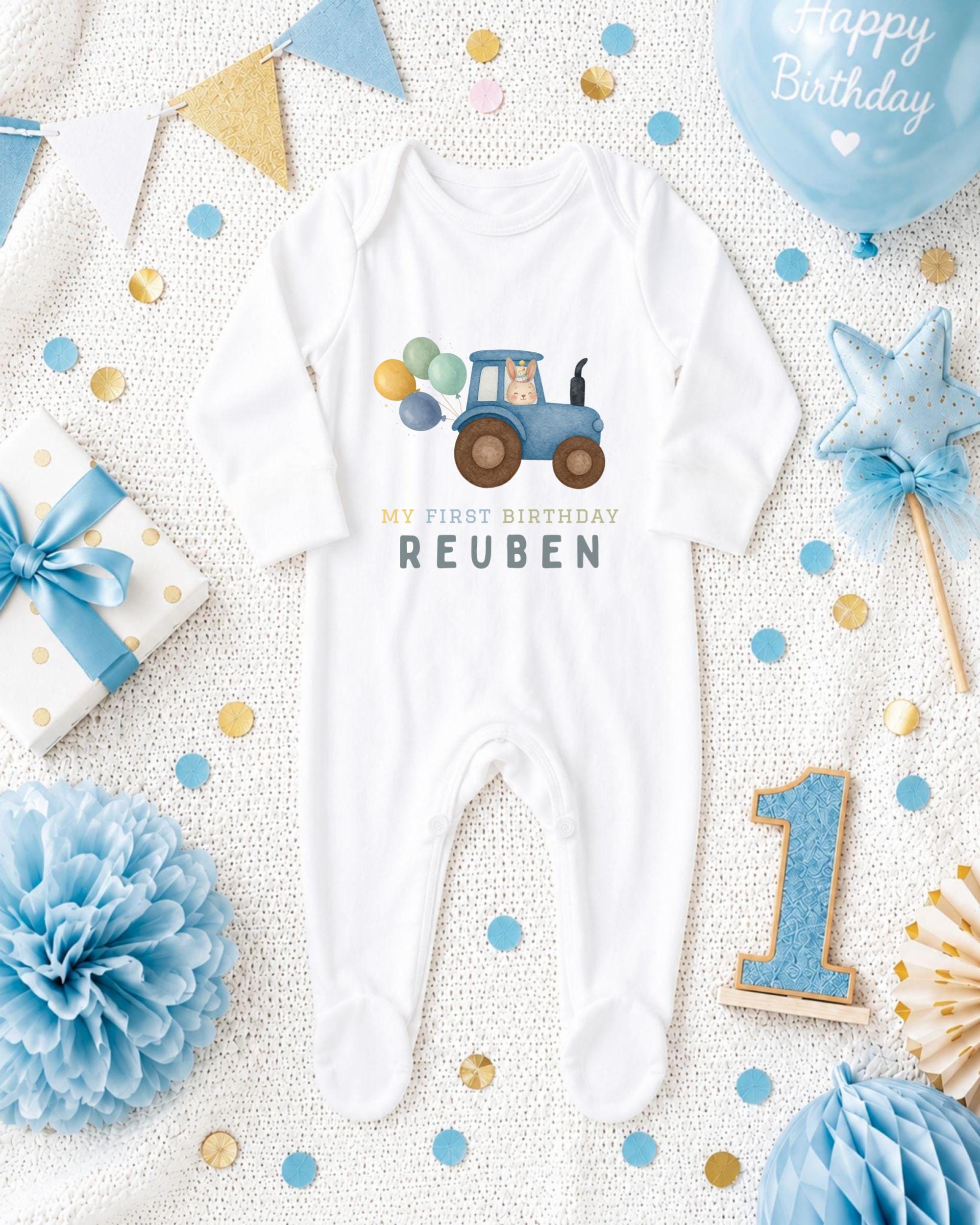 A baby onesie with a tractor design, surrounded by birthday decorations such as a blue balloon, a star, and confetti.