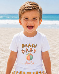 A young boy with blond hair is smiling and wearing a white t-shirt with the text "BEACH BABY Amelia" printed on it.