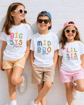 Three children wearing white t-shirts with colorful text and names printed on them, smiling and posing against a white brick wall.