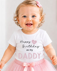A smiling baby girl wearing a white t-shirt with text and a pink bow, standing in front of a white background.
