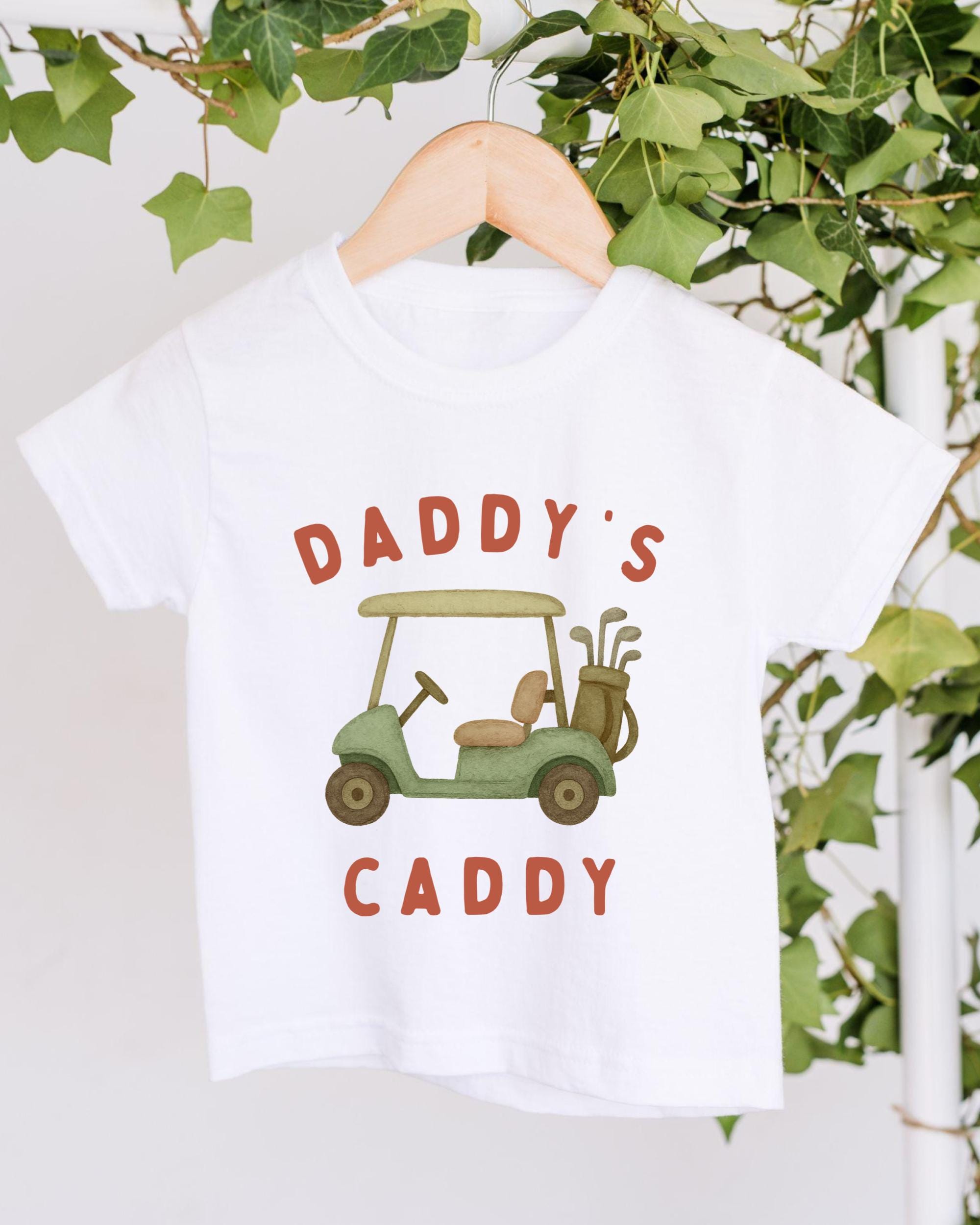 A white t-shirt with a graphic design of a golf cart and the text "DADDY'S CADDY" printed on it.