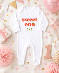 A white onesie with the text "Sweet on Eve" printed on it, surrounded by pink and gold confetti, a pink star, and a pink balloon.