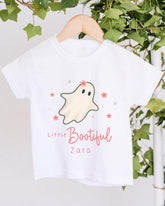 A white t-shirt with a ghost design and the text "Little Bootiful Zara" printed on it, hanging on a wooden hanger.