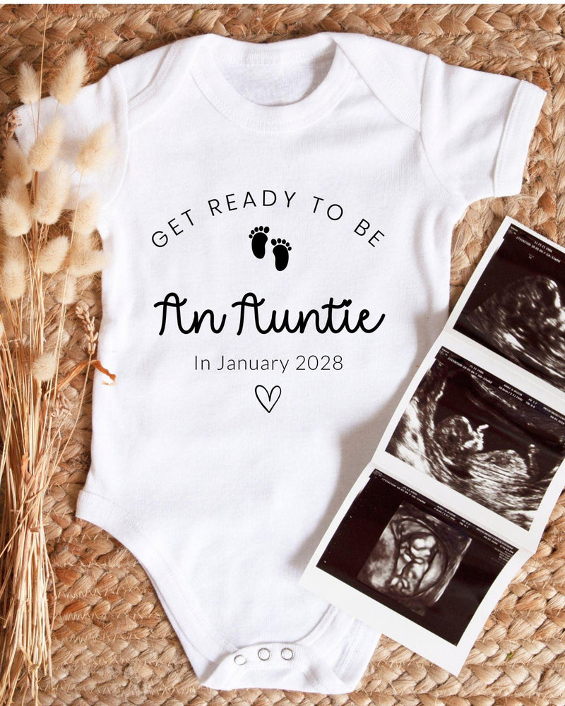 A white baby bodysuit with the text "Get ready to be An Auntie" and "In January 2028" printed on it, along with a heart symbol. The bodysuit is placed next to three black and white baby ultrasound images.