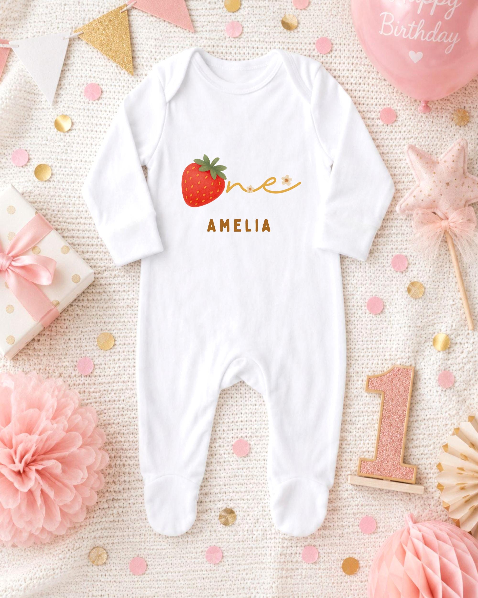 A white onesie with a red strawberry and the name "Amalia" printed on it, surrounded by pink and gold decorations, a pink balloon, and a pink flower.