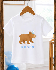 A white t-shirt with a cartoon bear wearing a party hat and the name "Wilson" printed on it, hanging on a wooden hanger.